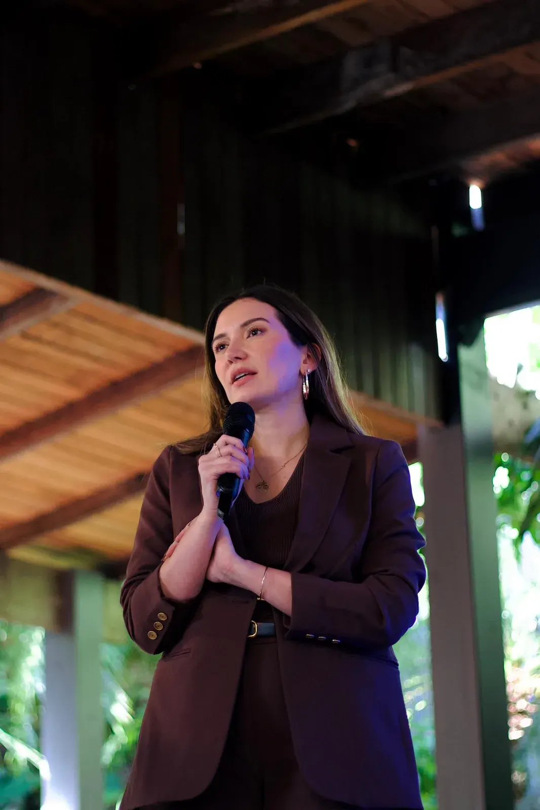 Apresentadora de blazer fala ao microfone sob a cobertura de madeira, com a vegetação do sítio ao fundo.