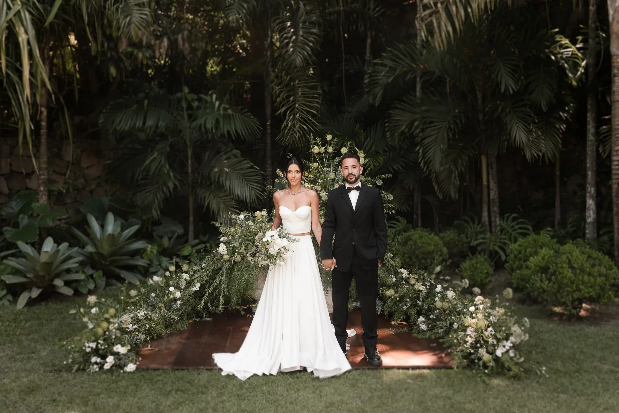 Everton Ribeiro, de smoking e gravata-borboleta, e Marília Nery, de corpete tomara que caia e saia longa branca, com um buquê de flores brancas, de mãos dadas sobre a passarela de madeira no gramado, diante de arranjos altos de flores brancas e folhagens, com as palmeiras e o muro de pedra ao fundo.