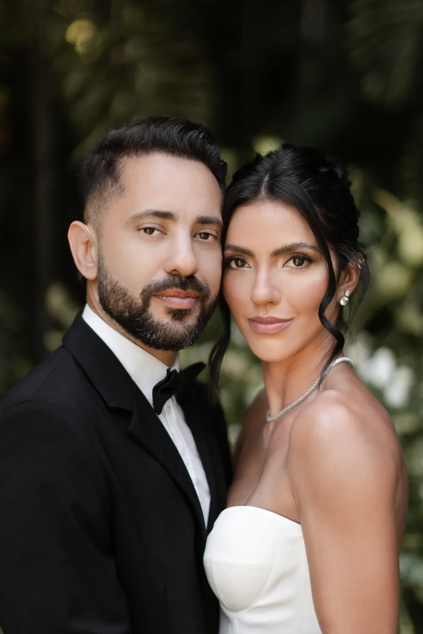 Retrato de Everton Ribeiro e Marília Nery, rosto com rosto, olhando para a câmera, com a folhagem do jardim desfocada ao fundo.
