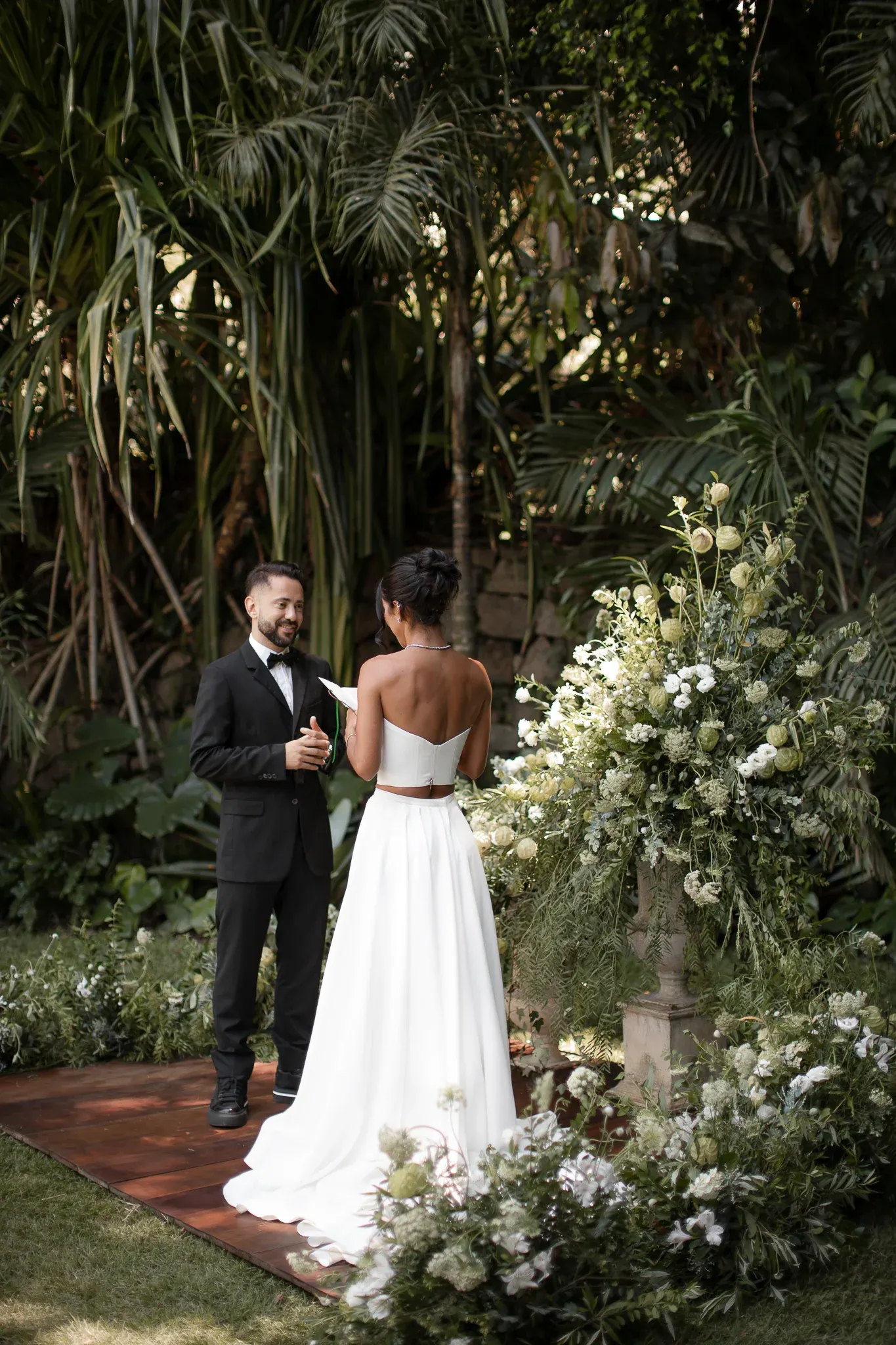 Marília Nery, de costas, lê os votos numa folha de papel diante de Everton Ribeiro, que sorri de mãos juntas, na passarela de madeira entre arranjos de flores brancas e verdes, sob as palmeiras.