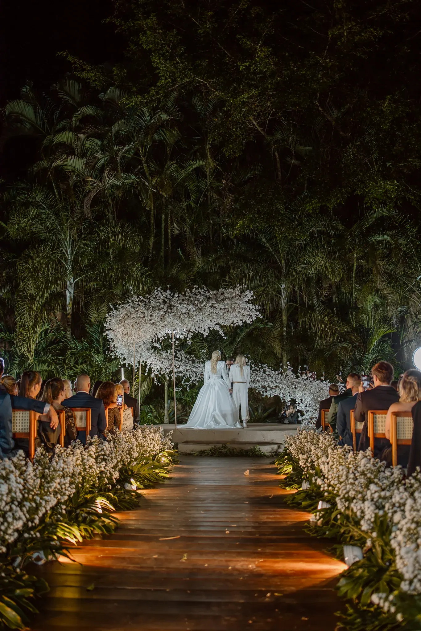 Cerimônia à noite: Monique Evans, de vestido branco de cauda longa, e Cacá Werneck, de terno branco, de mãos dadas no altar sob uma nuvem de flores brancas, diante das palmeiras iluminadas, com os convidados dos dois lados da passarela de madeira ladeada de mosquitinho.