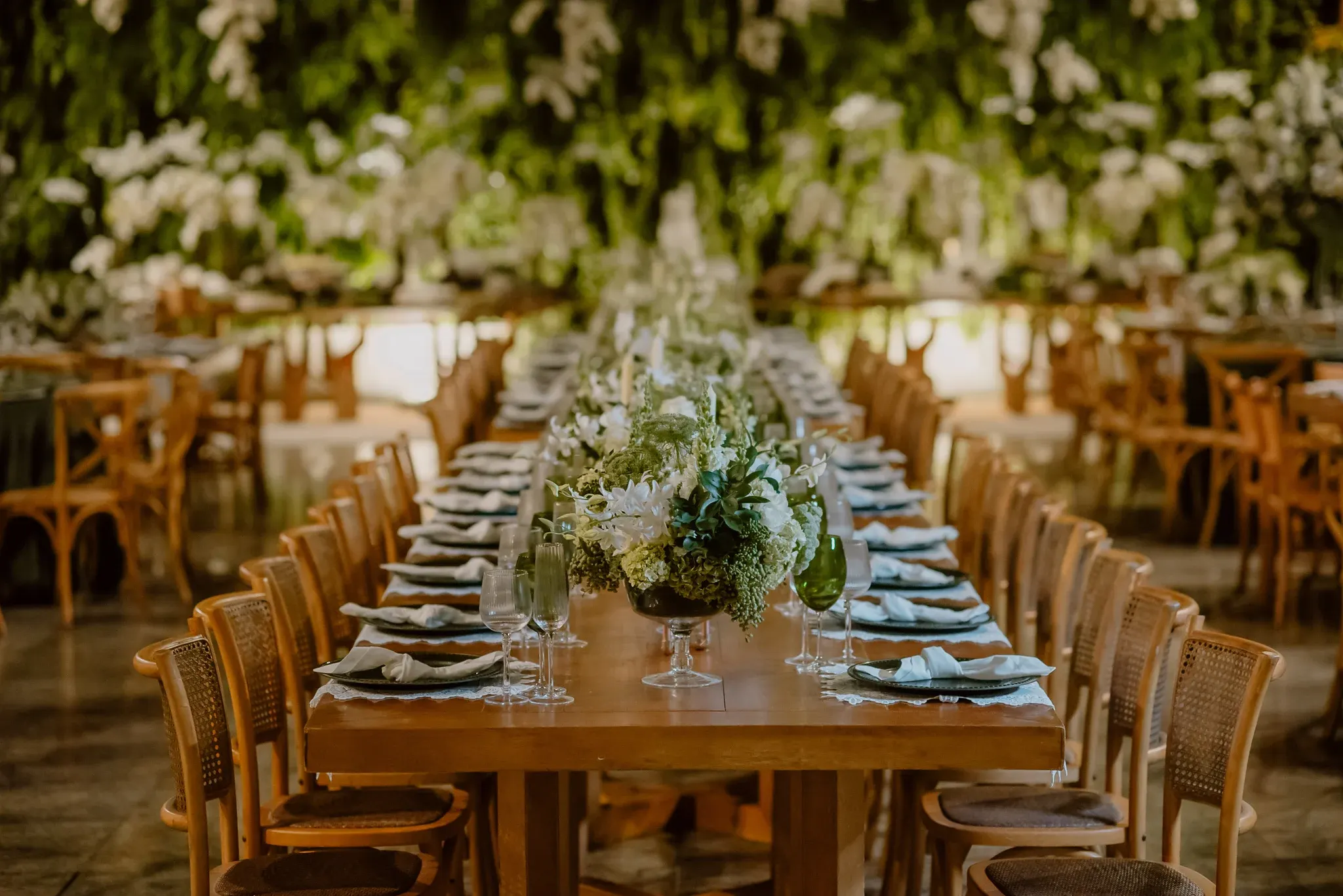 Mesa comprida de madeira posta para o jantar, com sousplats verde-escuros, guardanapos brancos com nó, taças verdes e de cristal e arranjos de flores brancas e folhagens, diante de uma parede de orquídeas brancas e verde.