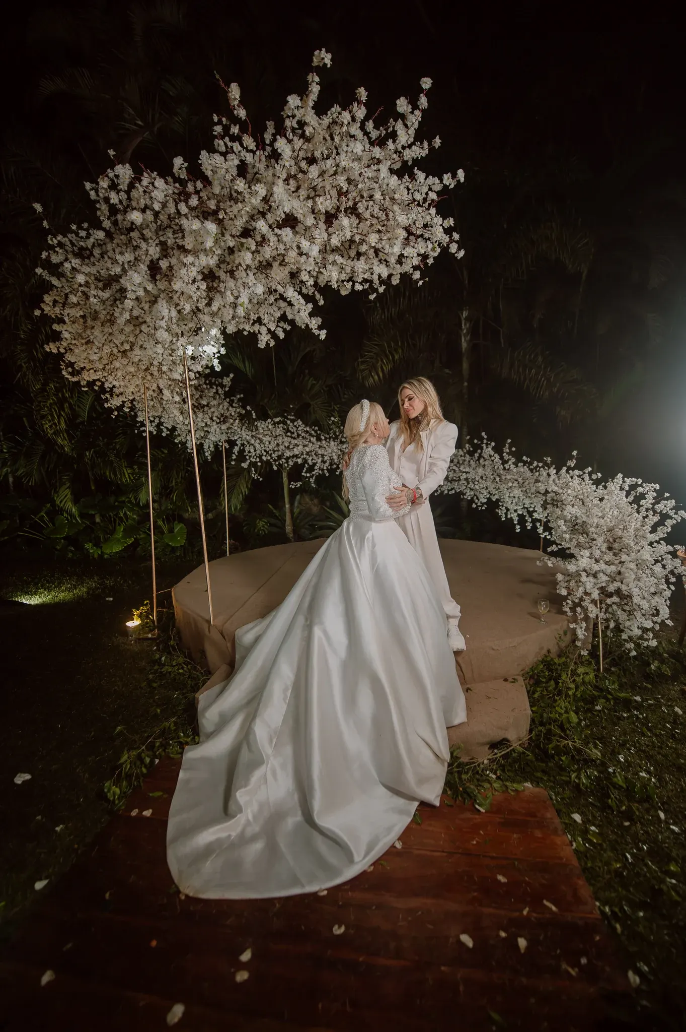 Monique Evans e Cacá Werneck abraçadas no altar redondo depois da cerimônia, à noite, sob o arco de flores brancas, com a cauda do vestido de cetim estendida sobre a passarela de madeira.