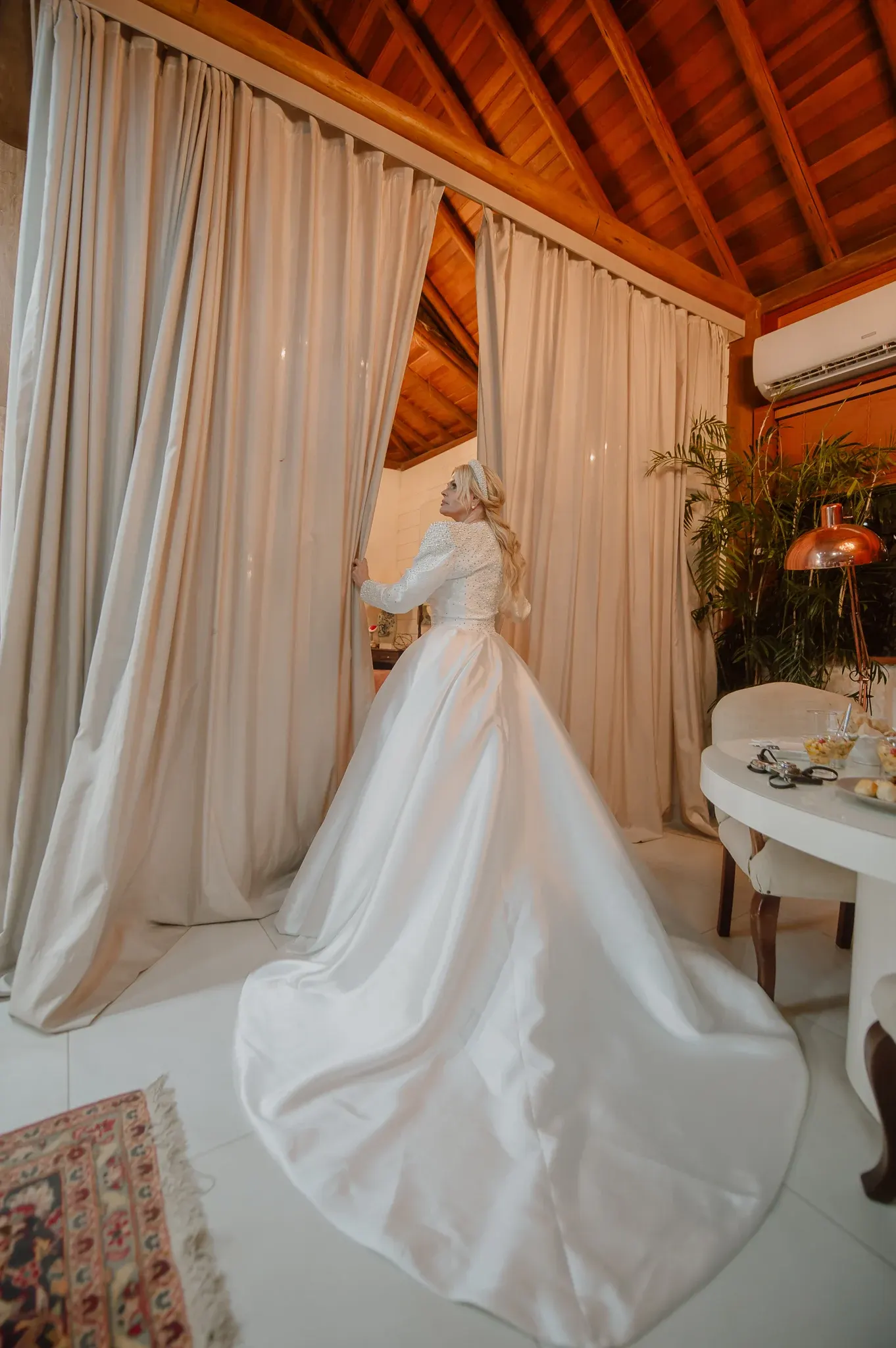 Monique Evans, de vestido de cetim com bolero bordado de pérolas e tiara, abre a cortina da suíte de preparação, de teto de madeira, com a cauda do vestido espalhada pelo chão.