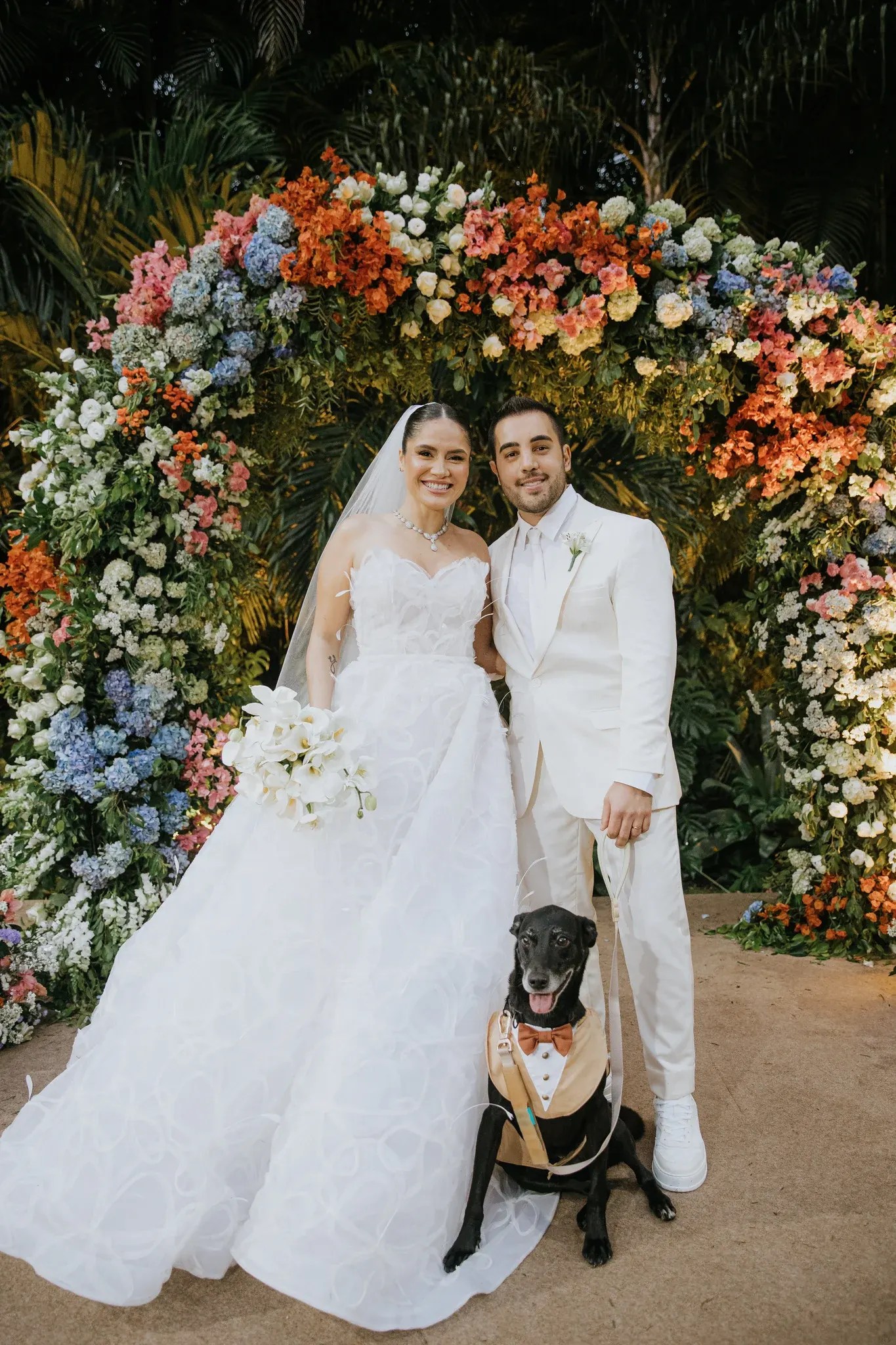 Joyce e Rodrigo sorriem sob o arco de flores depois da cerimônia, ela com um buquê de flores brancas e ele segurando a guia de Kaike, o cachorro preto do casal, de colete e gravata-borboleta cor de caramelo.