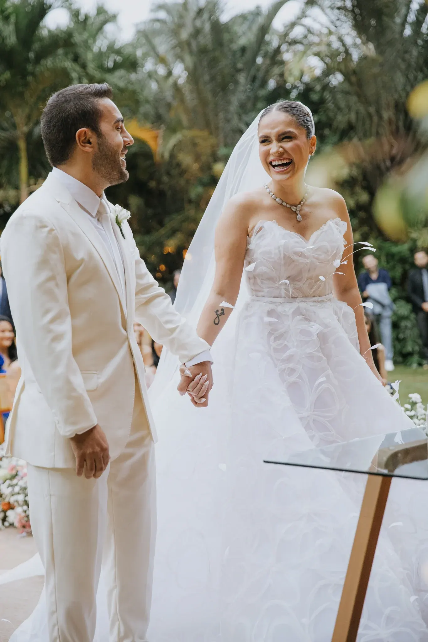 Rodrigo e Joyce de mãos dadas no altar, rindo, ela de véu e colar de brilhantes, com os convidados e as palmeiras do jardim ao fundo.