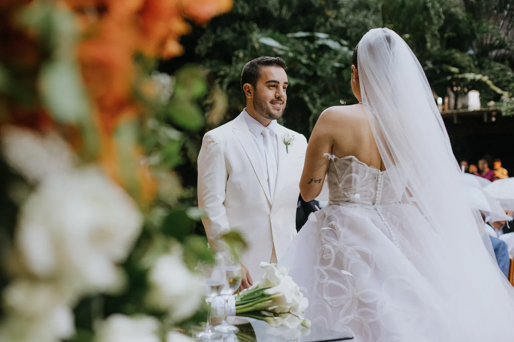 Rodrigo Melim, de terno branco e flor na lapela, sorri para Joyce durante os votos; ela está de costas, de véu longo e vestido tomara que caia bordado, diante de um buquê de copos-de-leite sobre a mesa de vidro, com a folhagem do jardim ao fundo.