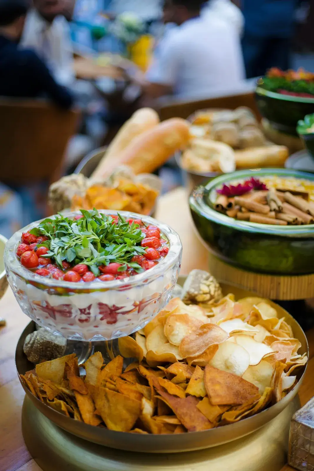 Mesa de ilha gastronômica montada em madeira, com travessa de tomates confitados e ervas, bandejas de chips e pães, e convidados sentados ao fundo, fora de foco.