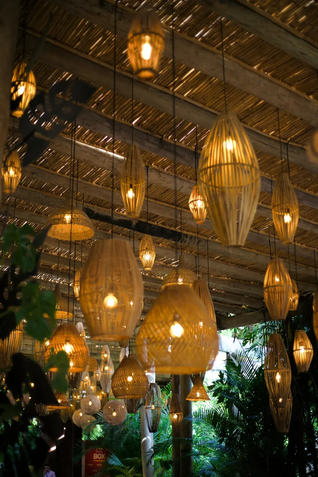 Luminárias de fibra de formatos diferentes penduradas sob o teto de palha, acesas ao entardecer.