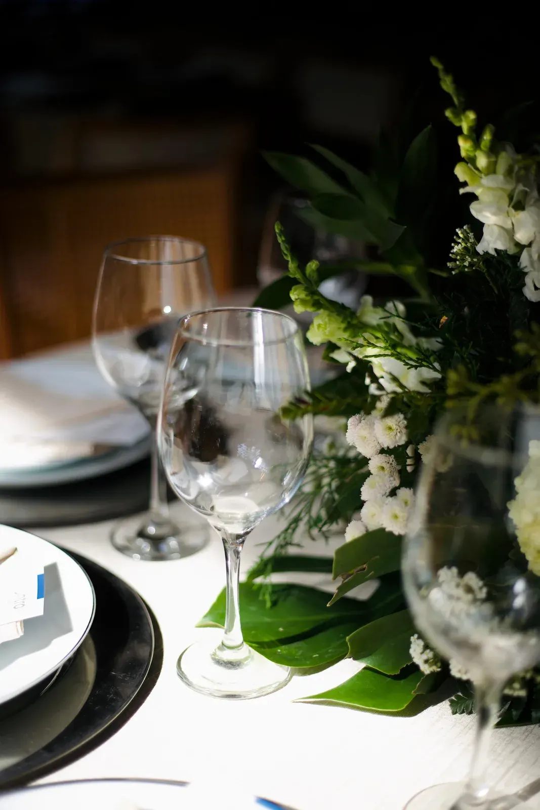 Lugar à mesa com taças de cristal, prato escuro sobre sousplat e arranjo de flores brancas com folhagem.