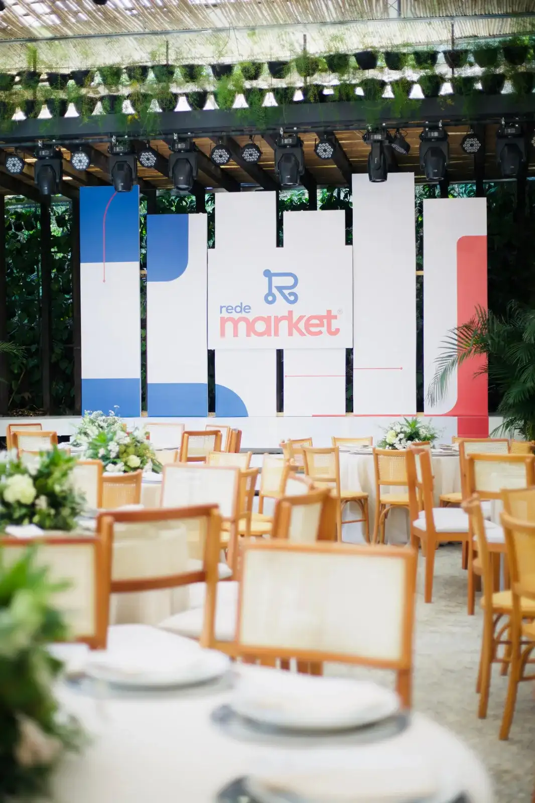 Salão montado para evento de empresa: mesas redondas com toalha branca e cadeiras de palhinha diante do painel cenográfico, sob a cobertura aberta para o jardim.