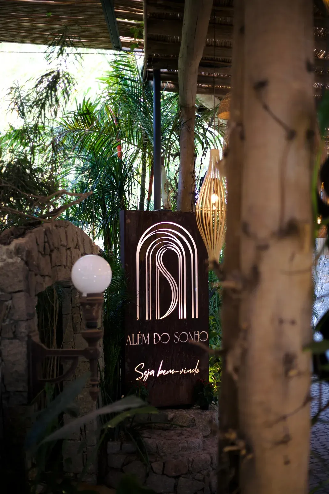 Totem de madeira com o símbolo do Além do Sonho e a inscrição Seja bem-vindo, iluminado à noite na entrada.