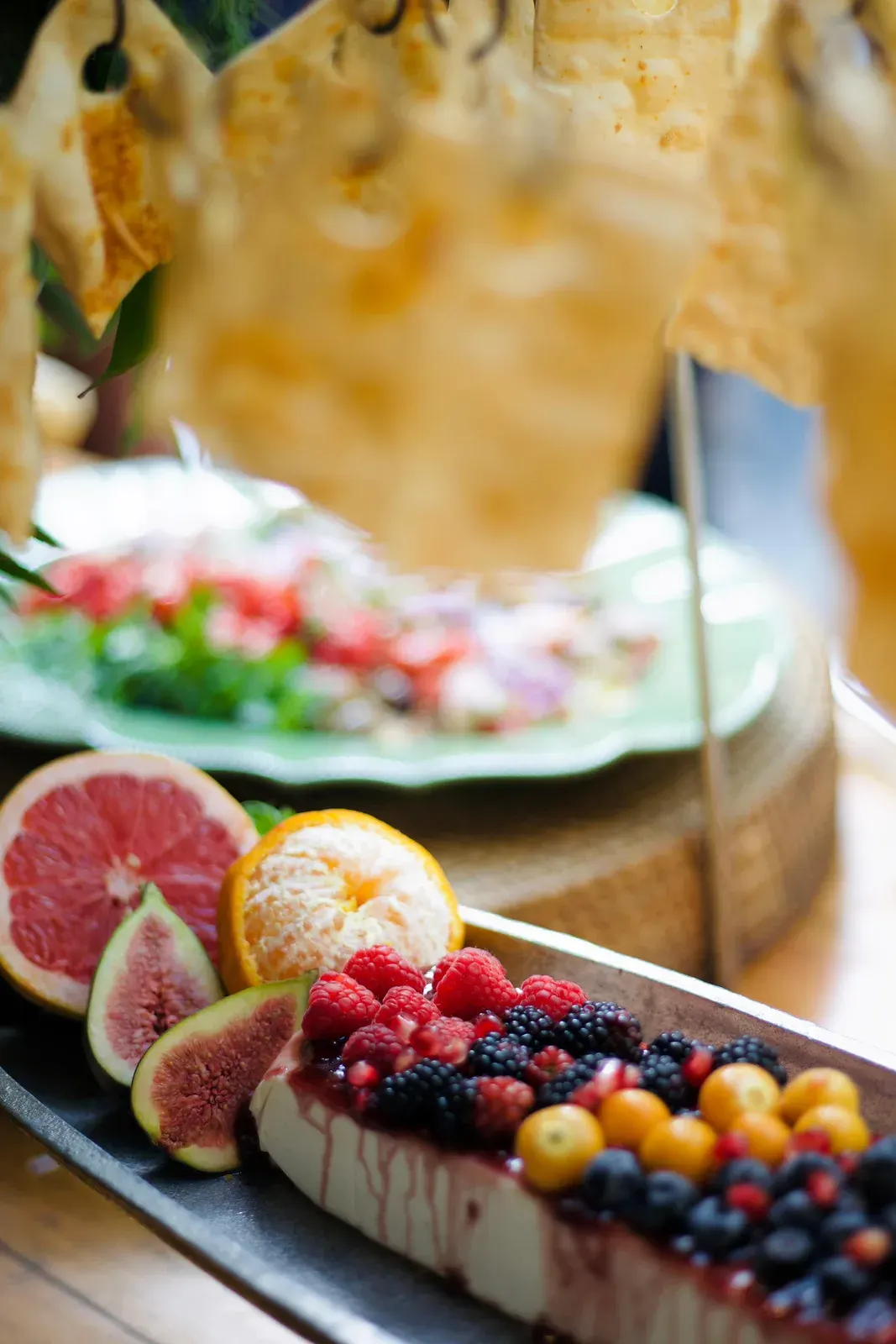 Travessa comprida com figos, frutas vermelhas, cítricos e queijos, montada sobre base de madeira.
