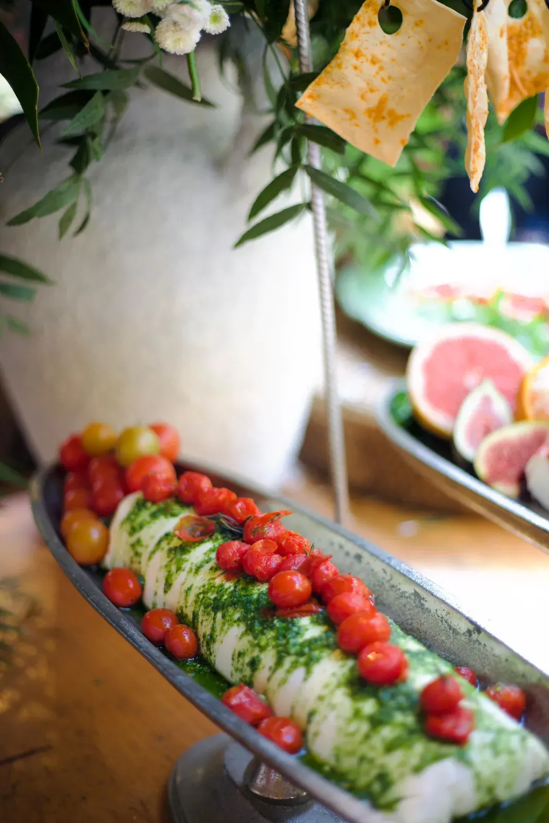 Travessa de tomates-cereja e ervas frescas sobre queijo fresco, com frutas ao fundo.